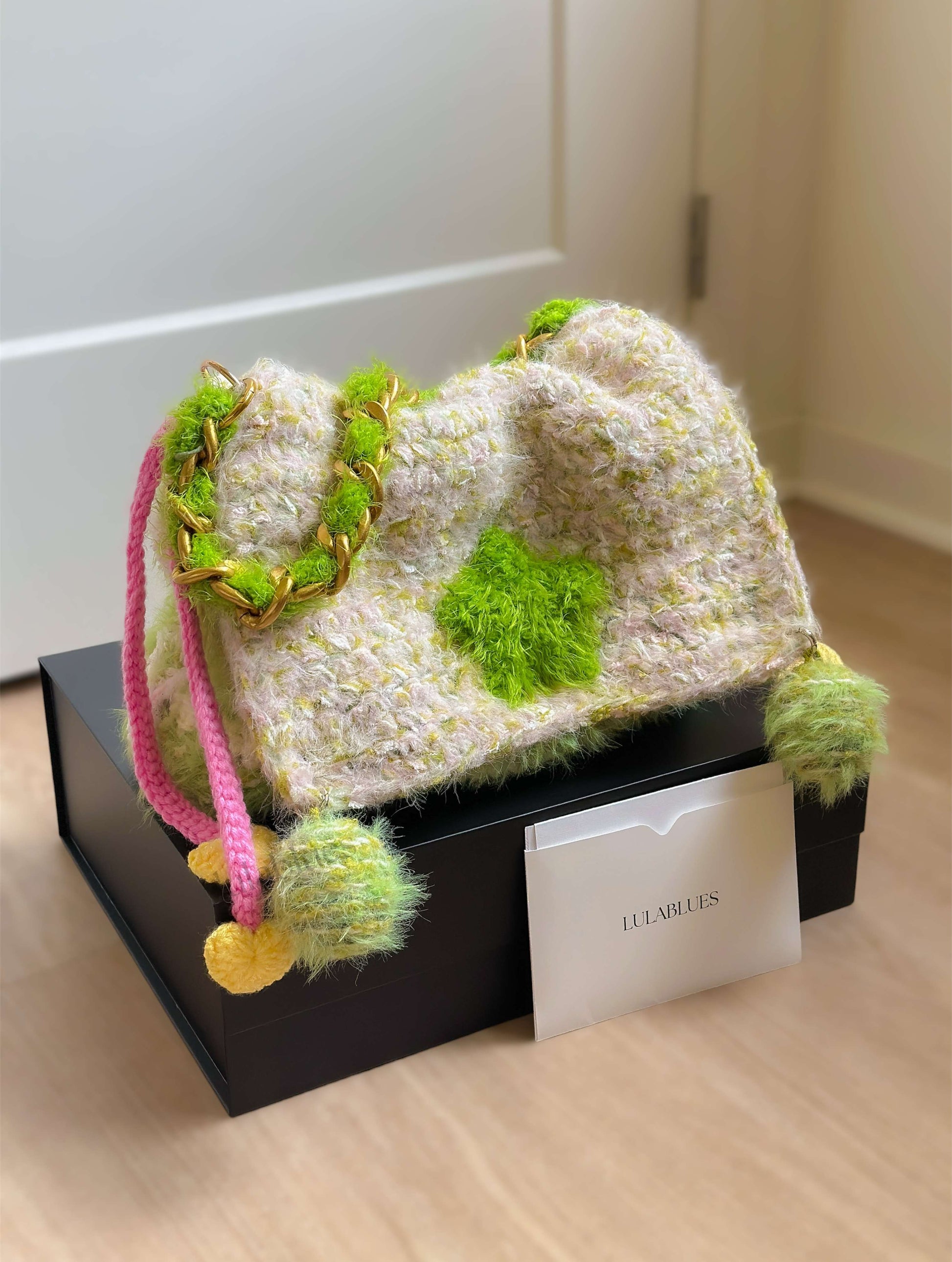 Textured handbag with green accents and pink strap on a black box, brand name visible